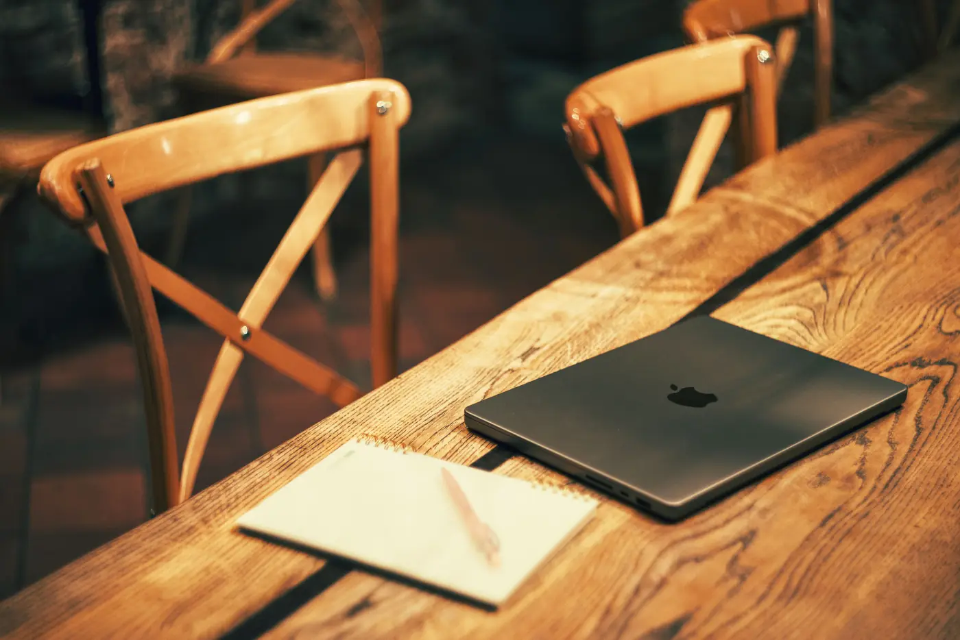 An open MacBook and a notebook with a pen on a warm wooden table