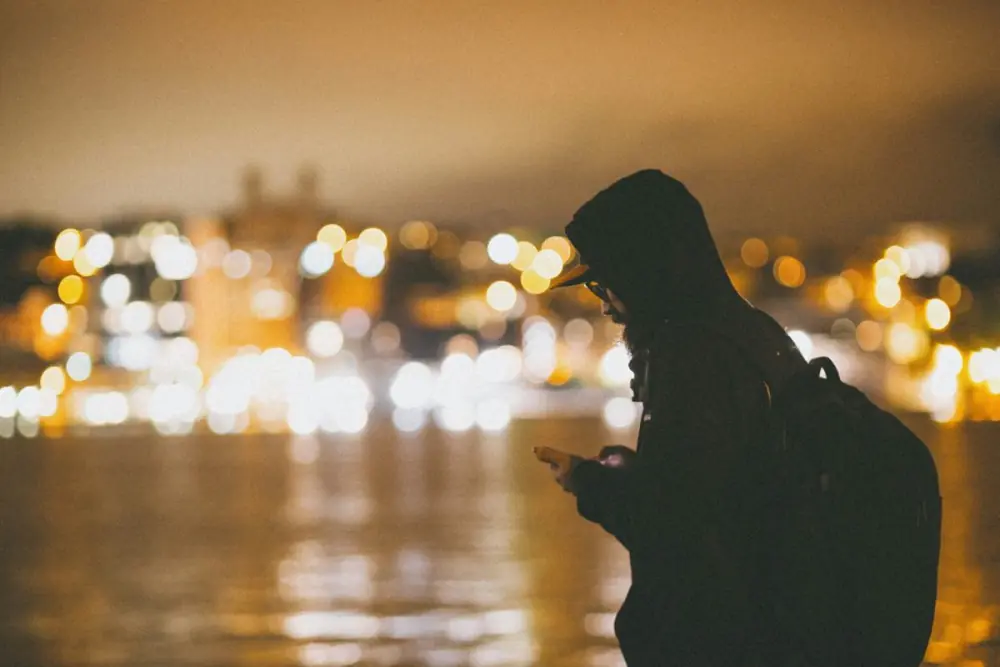 A hooded figure checking a phone at night against blurred city lights