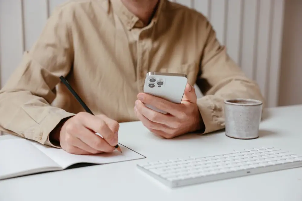 Hands writing in a notebook while holding an iPhone at a bright desk